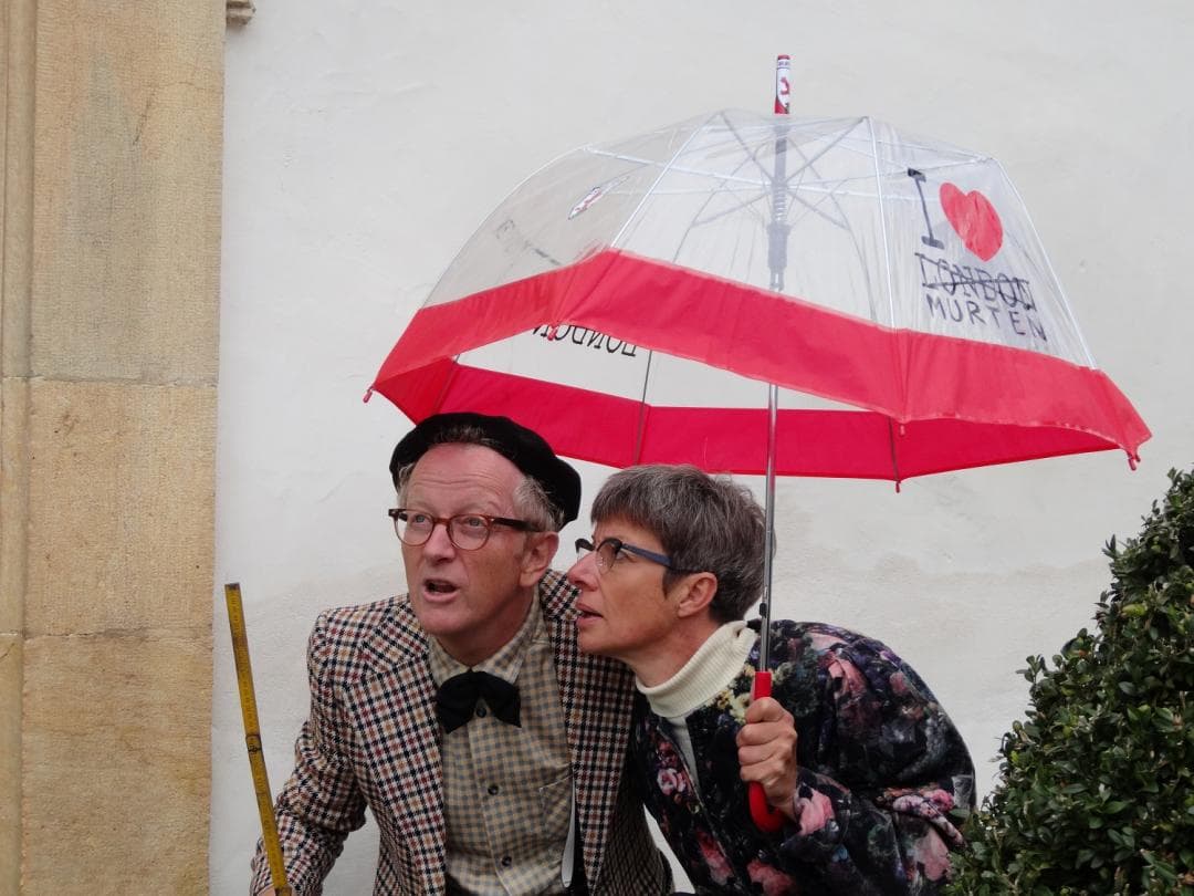 Amüsem Murten tour with humorous guides under an umbrella in front of a white wall.