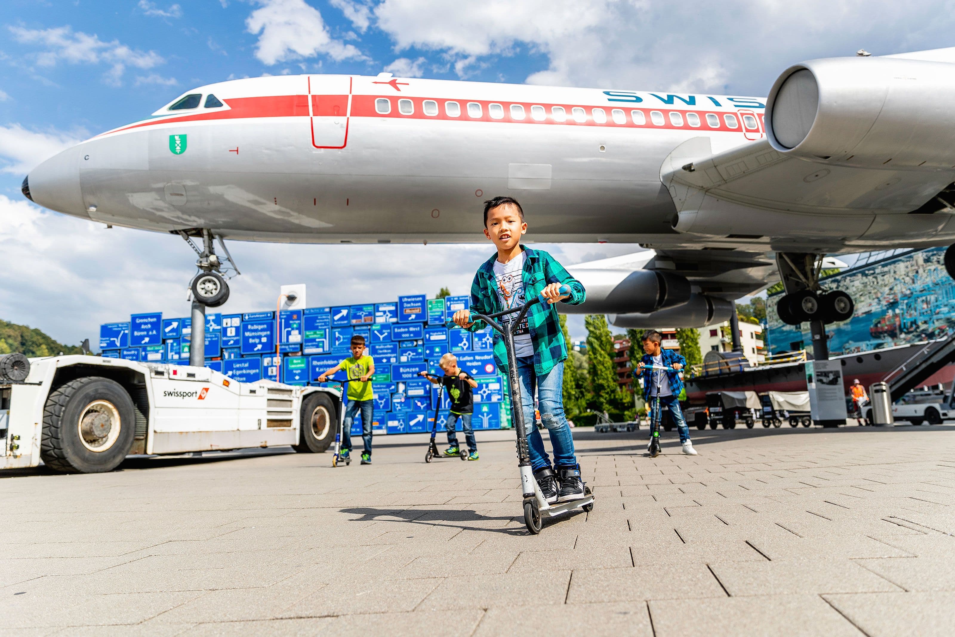 Children riding scooters at the Swiss Museum of Transport in Lucerne under an airplane